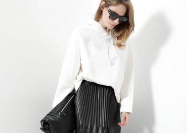 Eine Person mit Sonnenbrille und weißem Kapuzenpullover hält eine große schwarze Tasche. Sie trägt außerdem einen schwarzen Faltenrock. Der Hintergrund ist schlicht weiß.