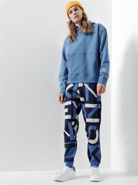 Eine Person mit einem blauen Kapuzenpullover, blau-schwarzen Hosen mit weißen Buchstaben und weißen Turnschuhen steht vor einem weißen Hintergrund. Sie hat lange Haare und trägt eine gelbe Beanie.
