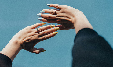 Zwei Hände mit bunten Nägeln, eine in Pastellblau und die andere in leuchtendem Gelb, heben sich vor einem klaren blauen Himmel ab. Die Hände tragen mehrere Ringe und der Hintergrund wirkt weich und verschwommen.