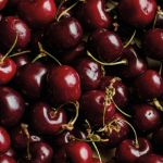 Eine Nahaufnahme zahlreicher reifer roter Kirschen mit grünen Stielen. Die Kirschen haben einen glänzenden Schimmer, der Frische signalisiert. Einige Stiele sind sichtbar und bilden einen Kontrast zu den satten Rottönen der Frucht.