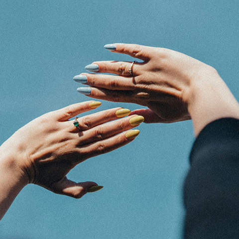 Zwei Hände mit bunten Nägeln strecken sich vor einem klaren blauen Himmel nacheinander aus. Eine Hand hat blaue Nägel, die andere gelbe. Sonnenlicht beleuchtet die Hände und an einem Finger befindet sich ein grüner Ring.