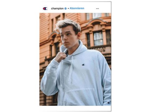 Eine Person mit einem hellblauen Kapuzenpullover posiert nachdenklich vor einem historischen Gebäude mit kunstvollen architektonischen Details.