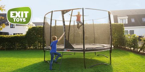 Zwei Kinder spielen auf einem runden Trampolin mit Sicherheitsnetz in einem Hinterhof. Ein Kind springt, während das andere eine kleine Leiter hochklettert. Die Szene spielt an einem sonnigen Tag mit Häusern im Hintergrund. In der Ecke ist das Logo von „Exit Toys“ zu sehen.