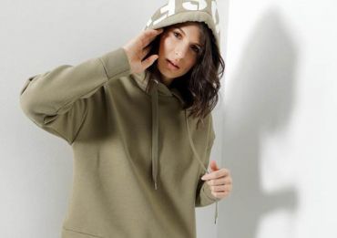 Eine Person mit langen braunen Haaren trägt einen olivgrünen Kapuzenpullover mit hochgezogener Kapuze und richtet ihn mit einer Hand. Sie steht vor einer hellen Wand und wirft einen Schatten darauf.