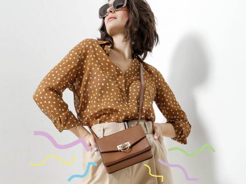Eine Person posiert selbstbewusst mit Sonnenbrille, einer braunen Bluse mit Tupfen, beigen Hosen und einer braunen Umhängetasche. Bunte Wellenlinien sorgen für eine kreative Note vor einem weißen Hintergrund.
