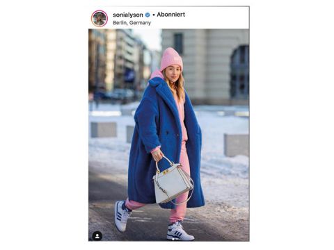 Eine Person läuft in einer verschneiten Stadtlandschaft. Sie trägt einen blauen Mantel, einen rosafarbenen Kapuzenpullover und eine dazu passende rosafarbene Mütze. Sie trägt eine weiße Handtasche und Adidas-Sneaker. Im Hintergrund sind Gebäude zu sehen.