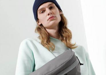 Eine Person mit langen, welligen Haaren trägt eine dunkle Beanie-Mütze und ein hellgrünes Sweatshirt. Sie hat einen neutralen Gesichtsausdruck und trägt eine graue Umhängetasche. Der Hintergrund ist eine minimalistische weiße Wand mit sanfter Beleuchtung.