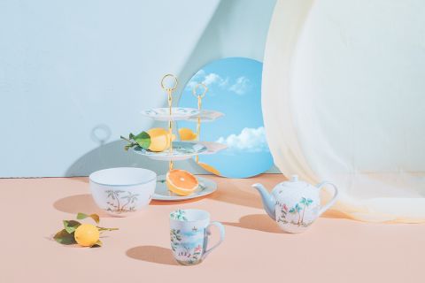 Ein pastellfarbenes Arrangement mit einem Dessertständer mit Zitronen, einer Teekanne, einer Teetasse und einer Schale mit Blumenmuster. Ein runder Spiegel reflektiert einen blauen Himmel mit Wolken in der minimalistischen Umgebung.