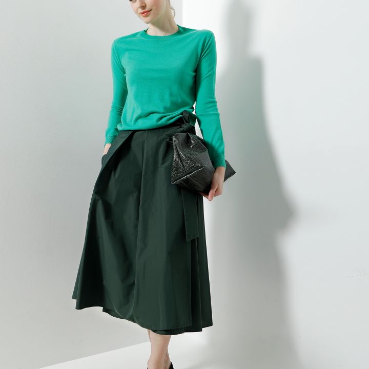 Eine Frau steht vor einer weißen Wand. Sie trägt ein grünes Langarmshirt, einen dunkelgrünen Rock, ein schwarzes Stirnband und schwarze Schuhe. Sie hält eine schwarze Tasche und lächelt sanft. Das Licht wirft einen weichen Schatten an die Wand.