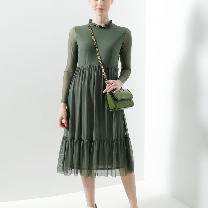 Eine Person trägt ein olivgrünes, langärmeliges, knielanges Kleid mit Rüschen am Ausschnitt und Saum, dazu weiße Turnschuhe und eine kleine grüne Umhängetasche. Der Hintergrund ist schlicht und weiß.