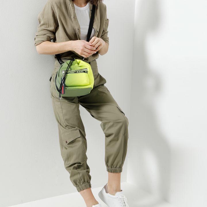 Eine Person mit schulterlangem Haar lehnt lächelnd an einer Wand. Sie trägt einen olivgrünen Overall, ein weißes T-Shirt und weiße Sneakers. Neben ihr hängt eine kleine neongrüne Umhängetasche.