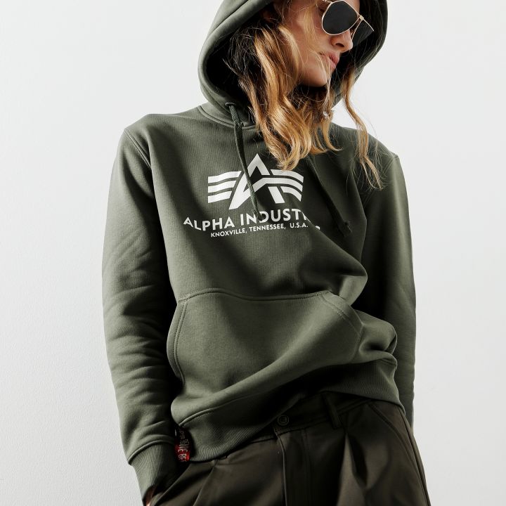 Eine Person mit dunkler Sonnenbrille, einem dunkelgrünen Kapuzenpullover mit dem Schriftzug „Alpha Industries“ und passender Hose posiert selbstbewusst vor einem schlichten Hintergrund. Ihre Hände stecken in den Kapuzenpullovertaschen und ihre Haare sind offen.
