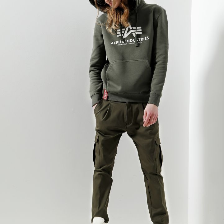 Person trägt einen grünen Kapuzenpullover mit „Alpha Industries“-Logo, grüne Cargohosen, Sonnenbrille und weiße Turnschuhe. Sie steht vor einer weißen Wand, die Hände in den Taschen, die Kapuze hochgezogen.