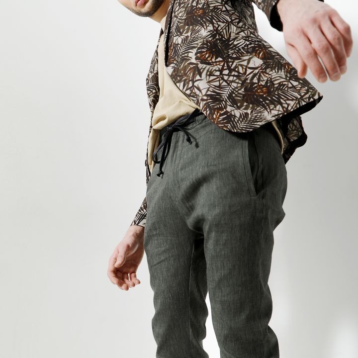 Eine Person mit kurzen dunklen Haaren posiert selbstbewusst vor einem weißen Hintergrund. Sie trägt eine gemusterte Jacke über einem beigen Hemd und einer olivgrünen Hose. Das Foto ist aus einem niedrigen Winkel aufgenommen, wodurch das Outfit betont wird.
