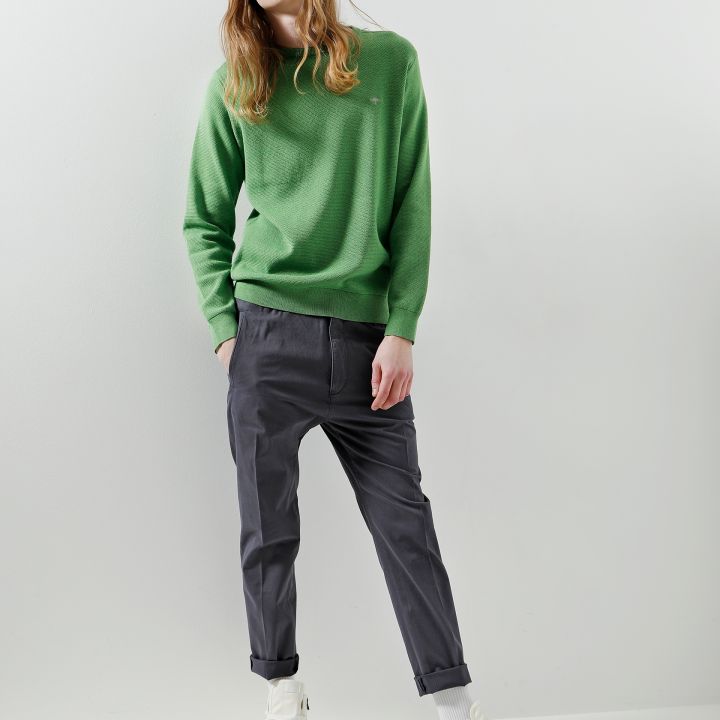 Eine Person mit langen Haaren, einer Sonnenbrille, einer marineblauen Mütze, einem grünen Pullover, grauen Hosen und weißen High-Top-Sneakers steht lässig vor einem schlichten weißen Hintergrund.