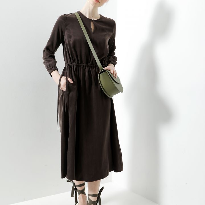 Eine Person steht in einem minimalistisch weißen Raum und trägt ein langes, dunkelbraunes Kleid mit taillierter Taille, gebundene Schuhe und eine grüne Umhängetasche. Die Person hat eine entspannte Pose und eine Hand in der Tasche.