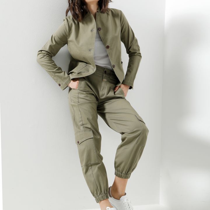 Eine Person in Khakijacke und -hose, weißem Hemd und Turnschuhen lehnt lässig an einer weißen Wand. Sie trägt eine Sonnenbrille und hat die Hände in den Taschen.
