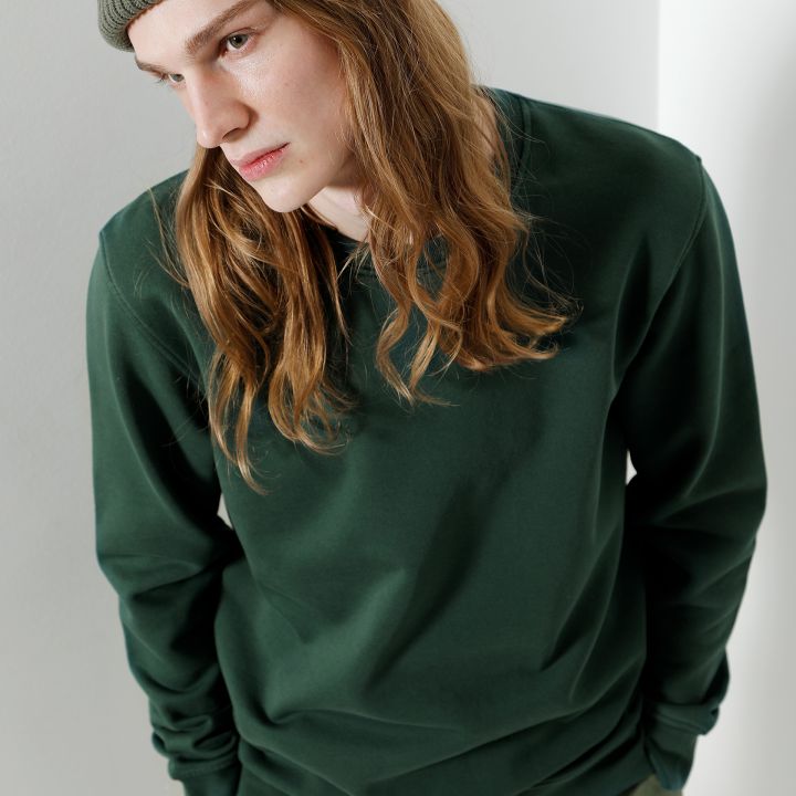 Eine Person mit langen Haaren, einer grünen Beaniemütze und einem grünen Sweatshirt steht vor einer hellen Wand. Sie hat den Kopf leicht geneigt und blickt zur Seite.