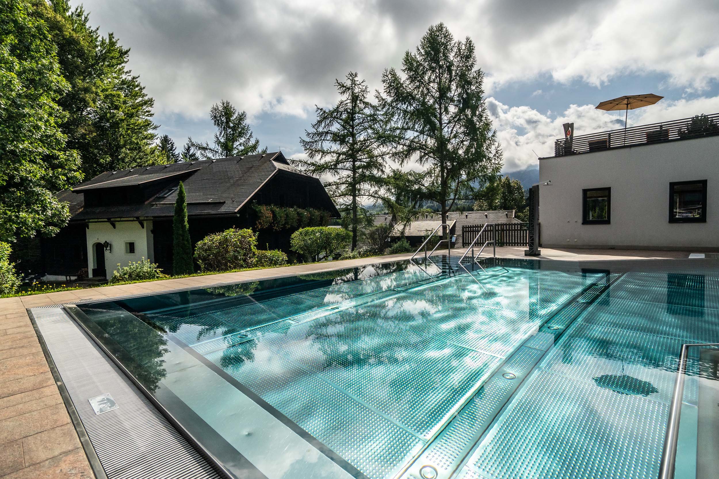 Ein moderner Außenpool mit Metallboden reflektiert das Sonnenlicht. Um den Pool herum stehen üppige Bäume und zwei Gebäude: ein traditionelles Holzhaus und ein weißes zeitgenössisches Gebäude. Wolken schweben am Himmel und tragen zur ruhigen Atmosphäre bei.