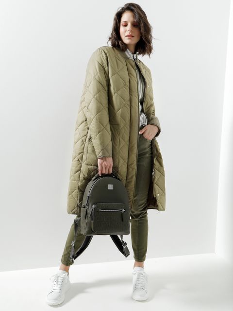 Eine Person, die einen langen, gesteppten olivgrünen Mantel über einem passenden Outfit trägt, posiert vor einem weißen Hintergrund. Sie hält einen kleinen grünen Rucksack und trägt weiße Turnschuhe.