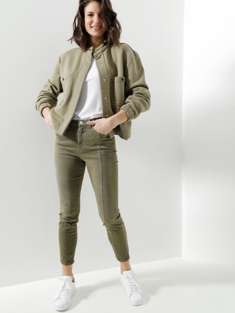 Eine Person steht in einem minimalistischen Raum und trägt eine Khakijacke, ein weißes T-Shirt, olivgrüne Röhrenjeans und weiße Turnschuhe. Sie hat eine entspannte Haltung, die Hände in den Taschen und lächelt in die Kamera.
