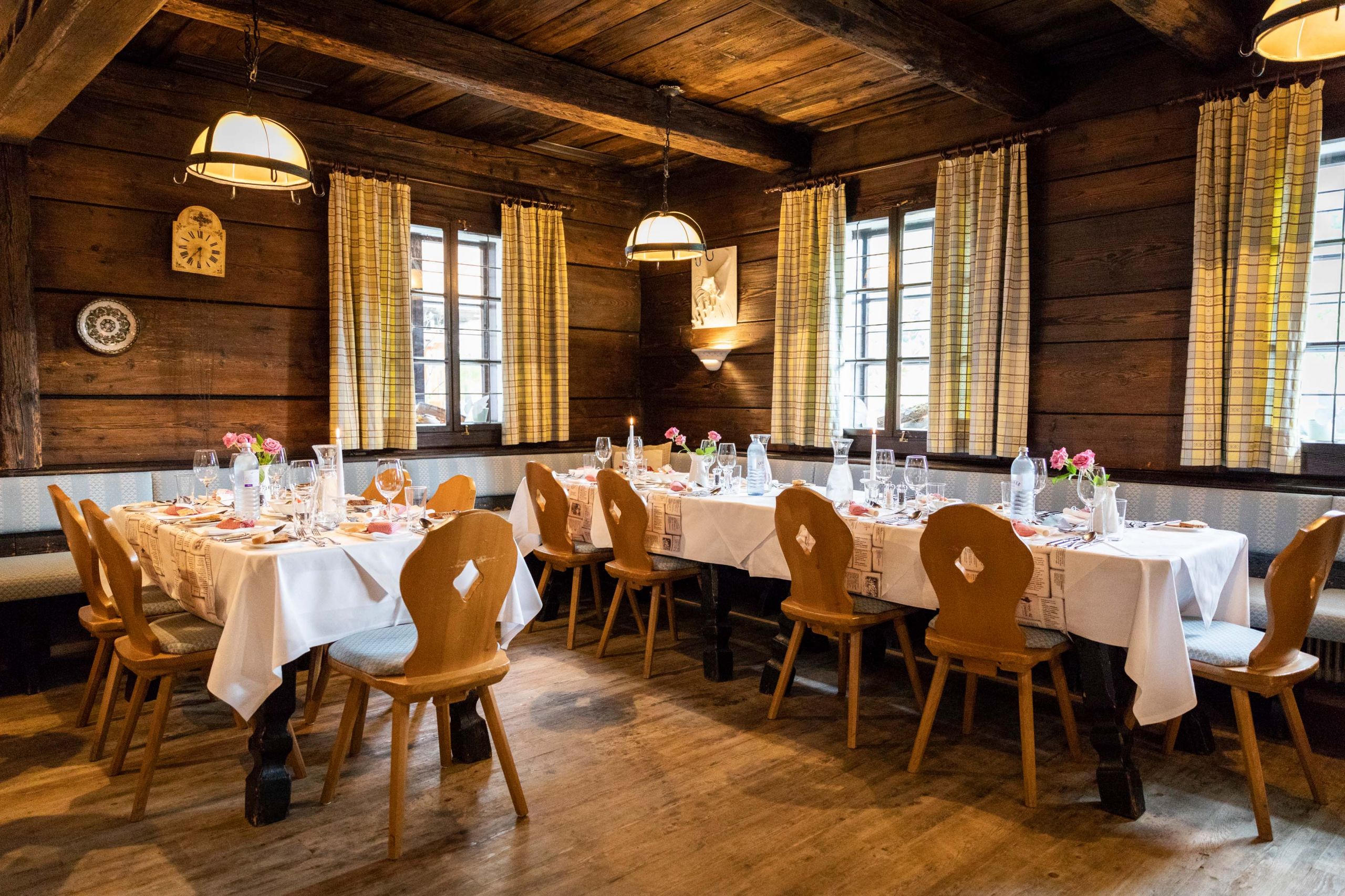 Ein rustikales Esszimmer mit Holzwänden und -decke, mit langen Tischen mit weißen Tischdecken, Glaswaren und Blumendekorationen. Holzstühle umgeben die Tische und große Fenster mit karierten Vorhängen lassen natürliches Licht in den Raum.
