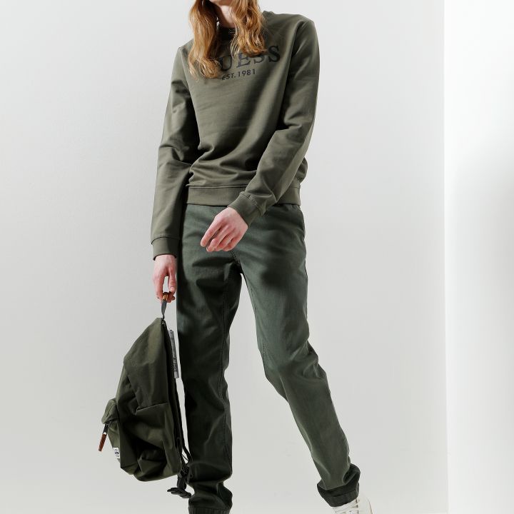 Person steht in minimalistischer Umgebung und trägt ein grünes Sweatshirt, eine passende Beanie und eine Hose. Sie hält einen grünen Rucksack in einer Hand und trägt weiße High-Top-Sneaker.