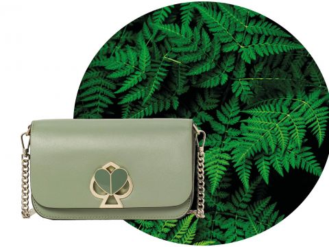 Eine grüne Handtasche mit einem goldenen Kettenriemen und einem dekorativen Verschluss ist vor einem Hintergrund aus leuchtend grünen Farnen positioniert.