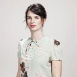Eine Frau mit zurückgebundenem dunklem Haar trägt eine hellgrüne Bluse mit Blumenmuster. Sie steht vor einem schlichten, hellen Hintergrund und blickt direkt in die Kamera. Die Bluse hat einen Spitzenbesatz und eine Schleife am Ausschnitt.