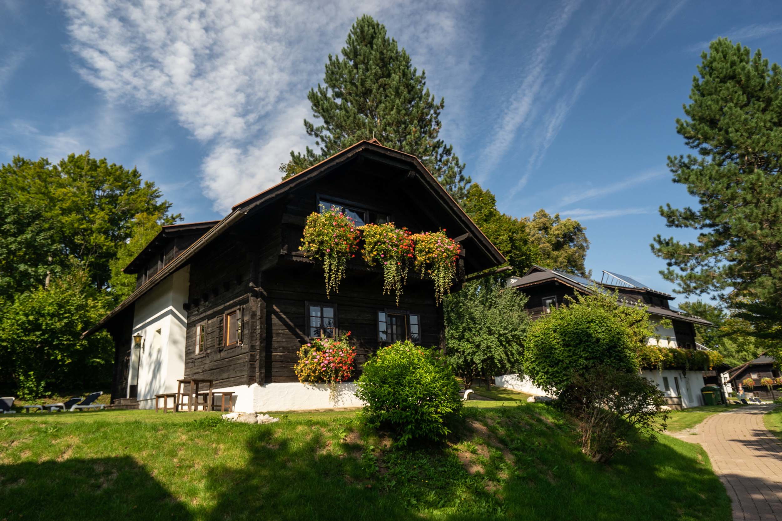 Ein rustikales Holzhaus mit Schrägdach ist von üppigem Grün und bunten Blumenkörben umgeben. Sonnenlicht wirft Schatten auf den Rasen, während hohe Bäume und ein klarer blauer Himmel mit dünnen Wolken die ruhige Szene vervollständigen.