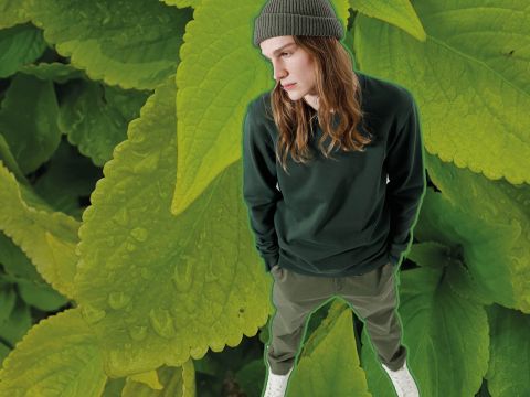 Eine Person mit langen Haaren, die eine grüne Mütze, einen grünen Pullover und Hosen trägt, steht vor einem Hintergrund aus großen, grünen Blättern mit Wassertropfen. Die Perspektive ist von oben und die Person blickt zur Seite.