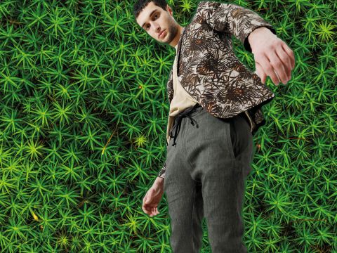 Eine Person in einer gemusterten Jacke und grauen Hosen posiert in einer Aufnahme aus niedriger Perspektive vor einem üppig grünen Blatthintergrund und schafft so eine lebendige und dynamische Komposition.