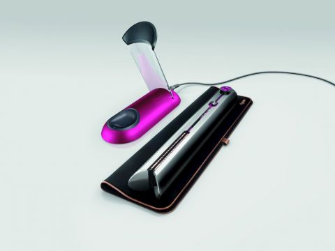 Ein eleganter, schwarzer Haarglätter mit silbernem Akzent auf einer weichen Hülle, neben einem rosa-schwarzen Haarstyling-Tool mit offenem Deckel, vor einem minimalistischen grauen Hintergrund.