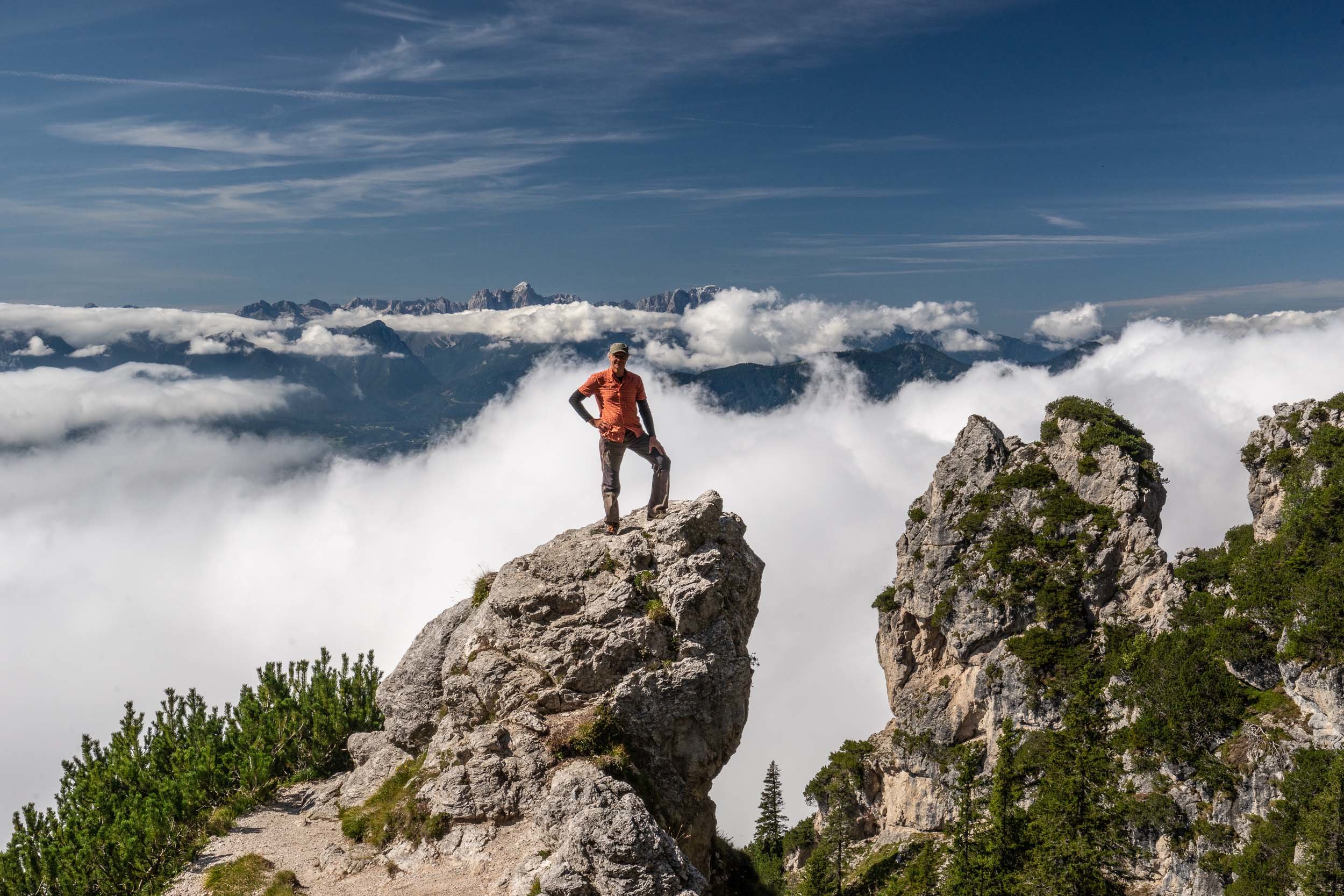 Eine Person steht triumphierend auf einer Felsklippe, umgeben von Wolken und einer weiten Berglandschaft. Der Himmel oben ist klar und blau und bildet einen Kontrast zu den weißen Wolken unten. Grüne Sträucher säumen das felsige Gelände.