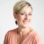 Eine lächelnde Person mit kurzen, blonden Haaren blickt in die Kamera. Sie trägt ein pfirsichfarbenes Oberteil und Perlenohrringe. Der Hintergrund ist schlicht weiß.