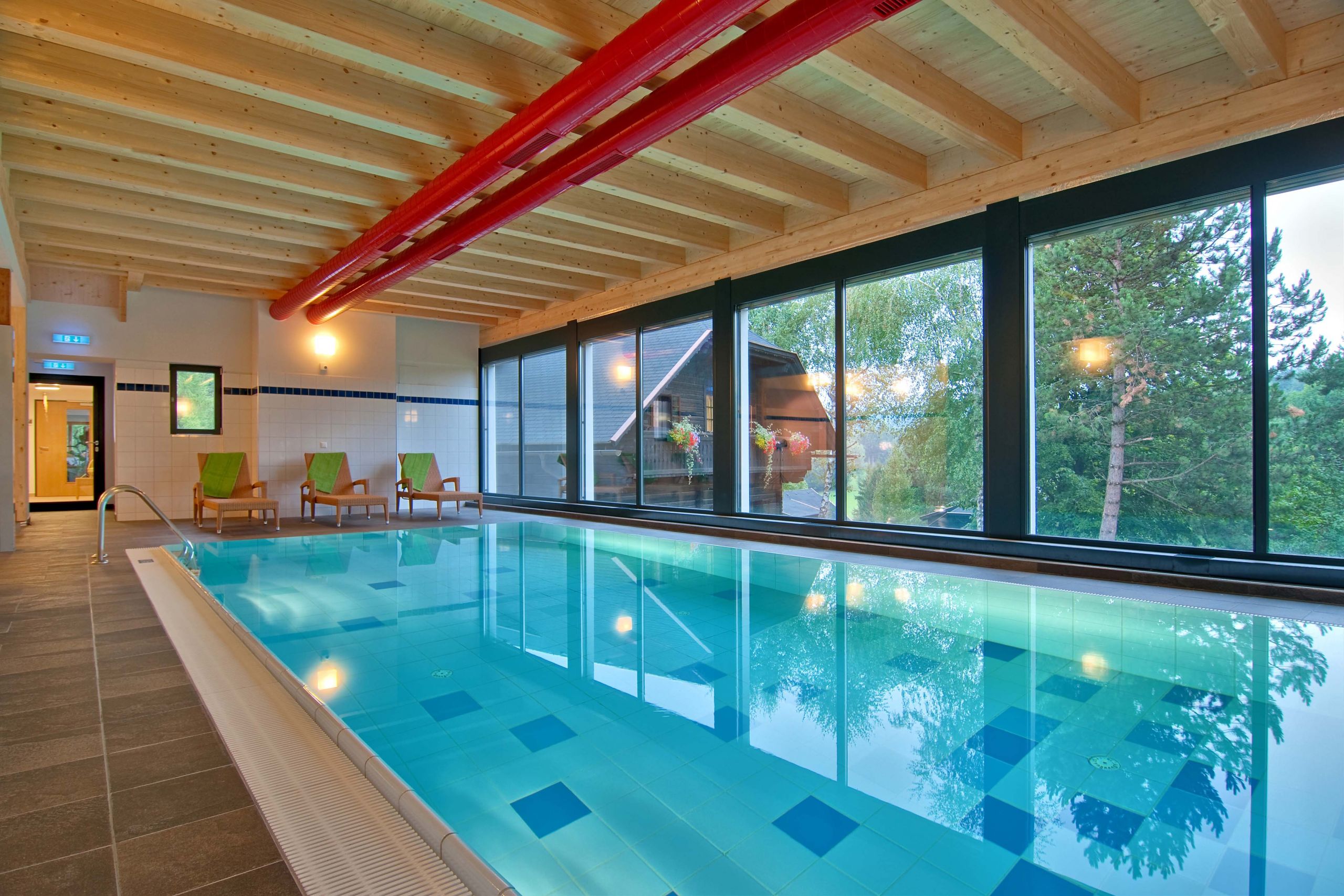 Innenpoolbereich mit großen Fenstern, die einen Blick auf die Bäume draußen bieten. Der Pool ist hellblau gefliest und auf einer Seite sind mehrere grüne Liegestühle aufgestellt. Die Decke besteht aus Holzbalken und roten Rohren.