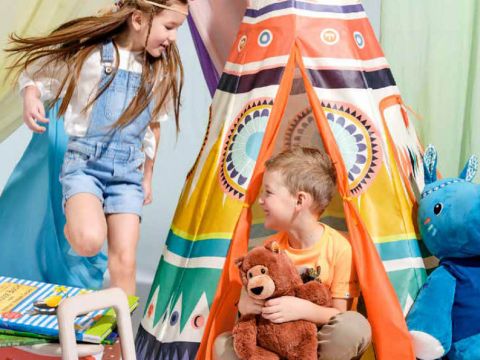 Zwei Kinder spielen in einem bunten Tipi. Ein Mädchen mit Stirnband und Jeans-Overall steht lächelnd draußen. Ein Junge drinnen hält einen Teddybären. In der Nähe sind bunte Bücher gestapelt und ein großes blaues Stofftier ist zu sehen.