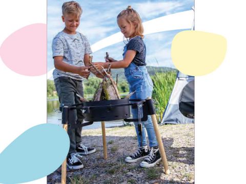 Zwei Kinder bauen draußen auf einem erhöhten Gestell ein kleines Gebilde aus Stöcken. Im Hintergrund sind ein Zelt und Grünpflanzen unter einem teilweise bewölkten Himmel zu sehen. Die Kinder lächeln und sind auf ihre Tätigkeit konzentriert.