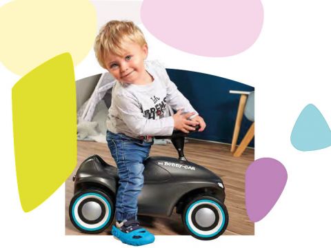 Ein Kleinkind mit blondem Haar, das ein weißes Langarmshirt und blaue Jeans trägt, fährt in einem schwarz-weißen Spielzeugauto durch einen Raum. Im Hintergrund sind bunte abstrakte Formen zu sehen.
