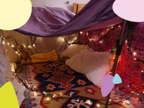 Eine gemütliche Deckenburg mit funkelnden Lichterketten, Kissen und Stofftieren im Inneren. Die Burg ist mit einer lila Decke bedeckt und steht auf einem gemusterten Teppich, was eine warme und einladende Atmosphäre schafft.