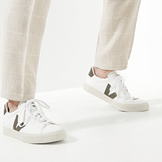 Person trägt beige karierte Hosen und weiße Turnschuhe mit dunkelgrünen Details und läuft auf hellem Untergrund.