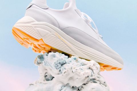 Ein stylischer Sneaker in Weiß- und Hellgrau-Design mit dicker Sohle und orangefarbenen Akzenten steht auf einem strukturierten, abstrakten weißen Sockel vor einem Himmelshintergrund mit Farbverlauf in Blau und Rosa.