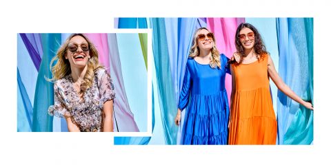 Zwei Frauen mit Sonnenbrillen, eine in einem blauen Kleid, die andere in einem orangefarbenen, lachen vor einem farbenfrohen, fließenden Stoff. Ein Einschub zeigt eine lächelnde Frau in einem Kleid mit Blumenmuster. Helle Außenkulisse mit leuchtenden Farben.