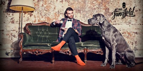 Ein Mann mit kariertem Jackett und Fliege sitzt auf einem Vintage-Sofa und hält eine Teetasse in der Hand. Er trägt orangefarbene Socken und sitzt einer großen Deutschen Dogge gegenüber. Die Umgebung hat eine rustikale, künstlerische Atmosphäre mit einer strukturierten Wand und einer eleganten Lampe.