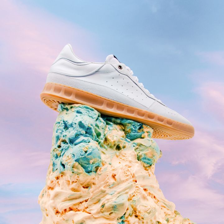 Ein weißer Turnschuh mit orangefarbener Sohle thront auf einem bunten, gewirbelten Eiscremehaufen vor der Kulisse eines pastellfarbenen Himmels mit rosa und blauen Farbtönen.