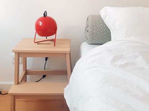 Ein ordentlich gemachtes Bett mit weißer Bettwäsche steht neben einem Nachttisch aus Holz. Auf dem Tisch steht eine kleine, runde, rote Lampe. Ein Kissen mit geometrischem Muster lehnt an der Wand neben dem Bett. Das Zimmer hat eine minimalistische Ästhetik.