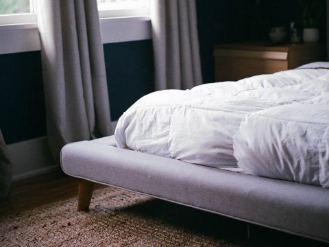 Ein ordentlich gemachtes Bett mit weißer Bettwäsche steht vor einer dunklen Wand. Licht fällt durch die Vorhänge und erhellt den Raum teilweise. Ein Nachttisch mit Pflanzen und ein gewebter Teppich sind sichtbar und schaffen eine gemütliche und einladende Atmosphäre.