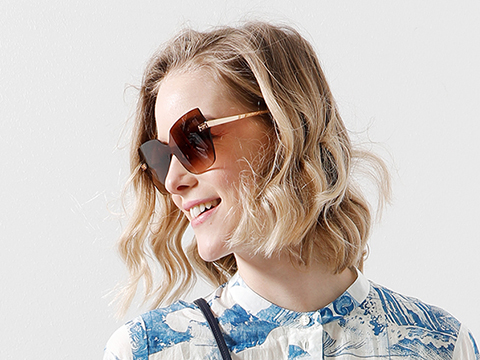 Eine Frau mit welligem blondem Haar, einer großen, quadratischen Sonnenbrille und einem gemusterten Hemd lächelt und blickt zur Seite vor einem schlichten weißen Hintergrund.