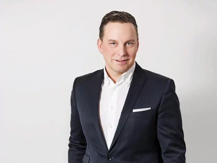 Ein Mann in schwarzem Anzug und weißem Hemd steht vor einem schlichten weißen Hintergrund. Er lächelt und hat die Hände in den Taschen.