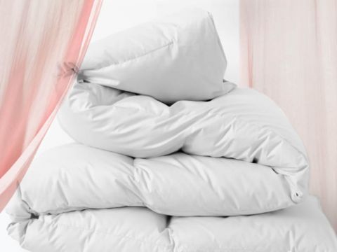 Ein Stapel weißer Kissen vor weißem Hintergrund, teilweise eingerahmt von durchsichtigen rosa Vorhängen an den Seiten. Die Kissen wirken plüschig und flauschig, lässig angeordnet.